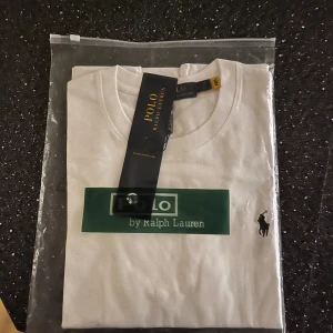Vit t-shirt från Polo Ralph Lauren - Vit t-shirt från Polo Ralph Lauren med klassisk grön logga framtill och broderad svart polospelare på bröstet. T-shirten har rund hals, korta ärmar och är gjord i mjuk bomull. Perfekt basic med snygg passform och stilren design.