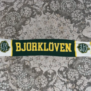Björklöven halsduk grön/gul/vit - Supporterhalsduk från Björklöven i grönt, gult och vitt med klubbens logga och texten BJÖRKLÖVEN. Klassisk design med fransar i ändarna och tydliga klubbfärger. Perfekt accessoar för att visa vilket lag du hejar på under matchen.