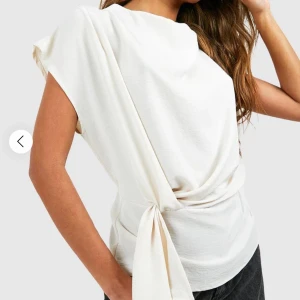 Vit omlottopp - Säljer en stilren vit omlottopp med knyt i midjan och korta ärmar. Toppen har en mjuk, lätt glansig yta och en avslappnad passform. Perfekt att styla med jeans eller kjol för en clean look.