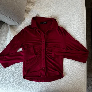 Vinröd skjorta med rynk från SHEIN - Snygg vinröd skjorta från SHEIN i storlek XS. Skjortan har långa ärmar, knappar framtill och cool rynkdetalj längs hela framsidan. Mjuk och stretchig känsla, perfekt för dig som gillar en trendig look med twist.