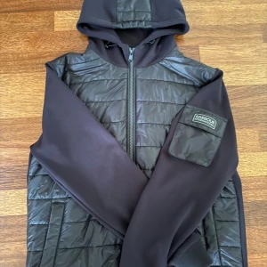 Svart quiltad Cardigan med huva från Barbour  - Svart dunväst från Barbour International med quiltad framsida, huva och dragkedja. Västen har släta ärmar i syntetmaterial och en ficka med Barbour-logga på ena ärmen. Perfekt för lager-på-lager och perfekt nu till hösten. Pris kan diskuteras