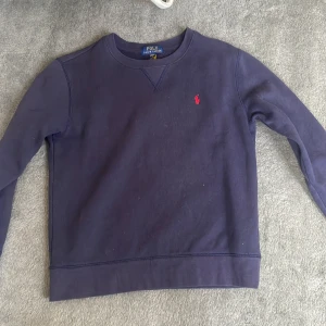 Mörkblå tröja från Polo Ralph Lauren - Mörkblå crewneck-tröja från Polo Ralph Lauren med klassisk röd broderad logga på bröstet. Tröjan har rund halsringning, långa ärmar och är tillverkad i mjuk bomull. Perfekt för en chill och stilren look.