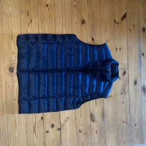 Mörkblå dunväst från Uniqlo - Säljer en mörkblå, ultralätt dunväst från Uniqlo. Västen har quiltad design, hög krage och dragkedja framtill. Perfekt för lager-på-lager och enkel att bära med sig tack vare den lätta känslan.