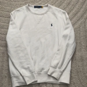 Vit sweatshirt från Polo Ralph Lauren - Vit sweatshirt från Polo Ralph Lauren med klassisk rund halsringning och broderad blå logga på bröstet. Tröjan har långa ärmar, ribbade muddar och är gjord i mjuk bomull. Enkel och stilren design som passar till allt.