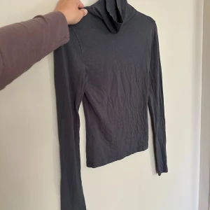 Mörkgrå polotröja i bomull - En stilren mörkgrå polotröja med lång ärm och tight passform. Tillverkad i mjuk bomull, perfekt att bära under jackor eller som lager på lager. Enkel och clean design utan detaljer, passar till allt.