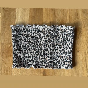 Leopardmönstrad bandeau tubtopp - Säljer en bandeau tubtopp med leopardmönster i svart och beige. Toppen har smockad struktur och vågiga kanter upptill och nedtill för en extra söt touch. Perfekt att styla med jeans eller kjol för en trendig look.