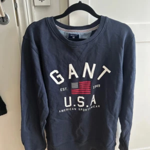 Mörkblå sweatshirt från GANT, S - Mörkblå sweatshirt från GANT med vit och röd USA-flagga och tryck framtill. Klassisk rund halsringning, långa ärmar och ribbade muddar. Tillverkad i 100% bomull för en skön och avslappnad känsla.