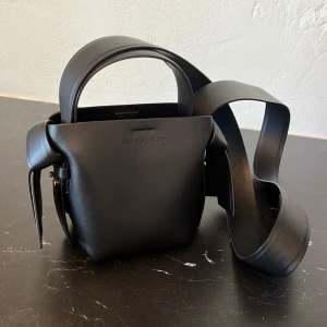 Acne Studios Musubi Micro tote  - Minimalistisk svart handväska från Acne Studios i slätt skinn. Väskan har en boxig form, bred axelrem och subtil logga framtill. Stängs med magnetknapp och har en innerficka. Perfekt för dig som gillar stilren design och vill ha en väska som sticker ut.