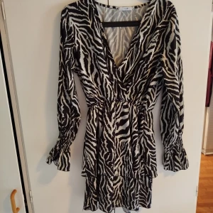 Zebramönstrad kortklänning från Holly & Whyte - Snygg kortklänning med zebramönster i svart och vitt från Holly & Whyte. Klänningen har v-ringning, långa ärmar med smock vid ärmslut och volangdetaljer i kjolen. Perfekt för dig som vill sticka ut med en trendig look. Storlek M. Funkar även för en mindre L