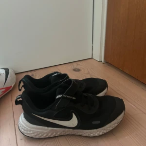 Svarta Nike Revolution sneakers - Svarta Nike Revolution sneakers med vit sula och klassisk vit swoosh på sidan. Skorna har snörning och kardborreband för extra smidig passform. Ovandelen är i mesh och syntetmaterial för bra andningsförmåga och komfort. Perfekta för träning eller vardagsstil.