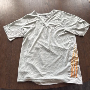 Ljusgrå Adidas t-shirt med gula ränder - Ljusgrå t-shirt från Adidas med klassiska gula ränder längs sidorna. Tillverkad i ett lätt och ventilerande polyestermaterial, perfekt för träning. T-shirten har korta ärmar och en sportig look med diskret Adidas-logga på bröstet.