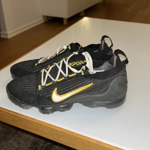 Nike Vapormax svart/gul sneakers - Nike Vapormax sneakers i svart med gula och vita detaljer. Ovandel i stickad mesh med vita snören och gula accenter. Swoosh-logga i vitt med gul kant. Synlig Vapormax-dämpning i sulan och blå Nike-logga bak. Perfekta för dig som gillar sportig och modern stil.