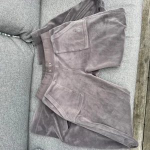 Gråa Juicy - Säljer ett par gråa Juciy med bred resår i midjan och stora fickor både fram och bak. Modellen är loose fit och har en avslappnad vibe, perfekt för chill dagar. Snygga detaljer med knappar på bakfickorna och diskret broderad logga framtill.