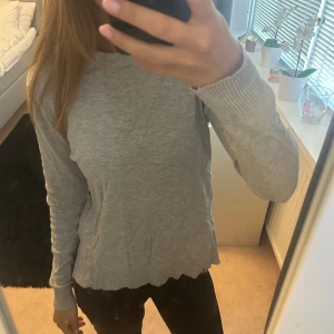 Grå stickad tröja med ribbade muddar - Mysig grå stickad tröja med rund halsringning och ribbade muddar vid ärmslut och nederkant. Tröjan har lång ärm och en klassisk, enkel design som passar perfekt till jeans eller leggings.