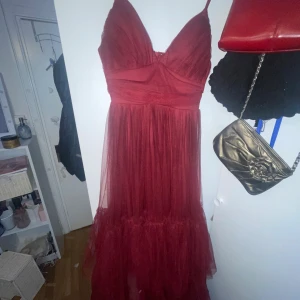 Vinröd långklänning i tyll - Säljer en vinröd långklänning med djup v-ringning och tunna axelband. Klänningen har flera lager tyll som ger en fluffig och romantisk känsla. Baktill finns en dragkedja för enkel påklädning. Perfekt för dig som vill sticka ut med en elegant look.