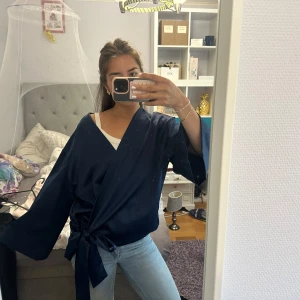 Mörkblå omlottblus med knytband - Säljer en mörkblå omlottblus med breda ärmar och knytband i midjan. Blusen har en snygg v-ringning och är tillverkad i ett glansigt, mjukt material som ger en lyxig känsla. Perfekt att styla med jeans eller kjol för en trendig look. Står ingen storlek men sitter så på mig med S
