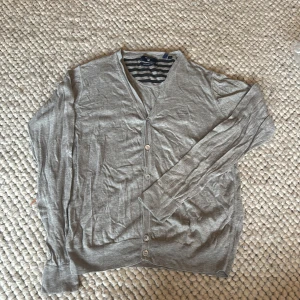 Grå kofta från GANT i S - Grå kofta från GANT med broderad logga på bröstet. V-ringad modell med knappar framtill och långa ärmar. Tillverkad i en lyxig mix av bomull, silke och ull. Perfekt för lager-på-lager och chill stil.