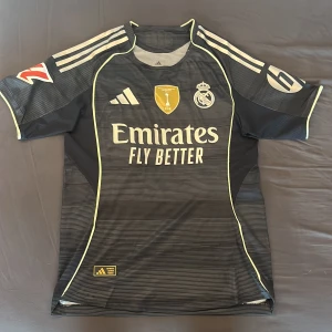 Real Madrid Huijsen 24 Adidas tröja - Snygg svart Real Madrid fotbollströja från Adidas med Huijsen 24 på ryggen. Tröjan har tunna horisontella ränder, gula detaljer och klubbmärke samt Adidas-logga broderade på bröstet. Tillverkad i lätt och ventilerande funktionsmaterial. Player Version