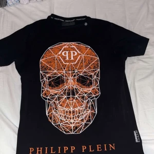 Philipp Plein t-shirt - Cool svart t-shirt från Philipp Plein med en stor grafisk döskalle i orange och vitt på framsidan. T-shirten har korta ärmar, rund hals och är gjord i mjuk bomull. Logotypen är tryckt i orange nedtill. Perfekt för dig som gillar edgy streetwear.