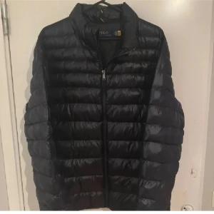  Polo Ralph Lauren puffer jacka  - Snygg svart pufferjacka från Polo Ralph Lauren i storlek M. Jackan har horisontella quiltade paneler, hög krage och dragkedja framtill. Materialet är glansigt och syntetiskt, perfekt för kalla dagar. Klassisk och stilren design som funkar till många outfits.