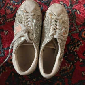 Glittriga sneakers med stjärndetalj - Säljer ett par sneakers med silverglittriga paneler och en ljusrosa stjärna på sidan. Skorna har snörning och rund tå, perfekt för dig som vill sticka ut lite extra. Bekväma och snygga till många outfits.