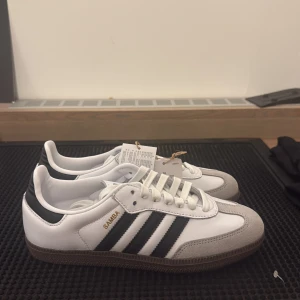 Adidas Samba OG vit/svart 37 1/3 - Adidas Samba OG sneakers i vitt skinn med svarta detaljer och klassiska tre ränder på sidan. Grå mockadetaljer vid tån och brun gummisula. Guldtexten 'SAMBA' på sidan och snörning framtill. Perfekt för dig som gillar retrostil och streetwear.( Äkta morsan jobbar där) Aldrig andvänd