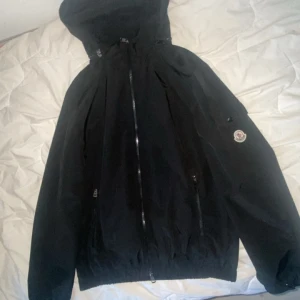 Svart vindjacka från Moncler - Svart vindjacka från Moncler med huva och dragkedja framtill. Jackan har Moncler-logga på ärmen, elastisk midja och ärmar samt praktiska fickor med dragkedja. Perfekt för dig som vill ha en clean och stilren look med exklusiv känsla.