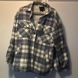 Rutig overshirt från 157 i grått och vitt - Overshirt från 157 i grått och vitt rutigt mönster. Jackan har klassisk krage, knäppning framtill med knappar och två bröstfickor med lock. Mjuk och lite tjockare känsla, perfekt för lager-på-lager. Passar dig som gillar avslappnad och trendig stil.