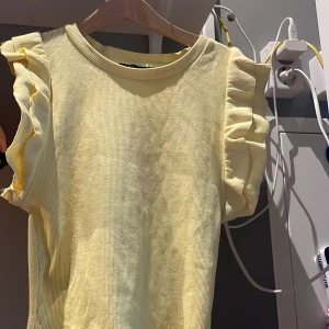Gul ribbad topp med volangärmar från Zara - Säljer en ljusgul ribbad topp med volangdetaljer på ärmarna. Toppen är ärmlös med rund halsringning och lätt croppad passform. 