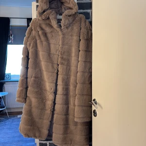 Beige pälsjacka med huva - Mysig och fluffig beige pälsjacka med huva och horisontella sömmar. Jackan är lång och har en oversized passform, perfekt för kalla dagar. Fodrad insida för extra värme och komfort.