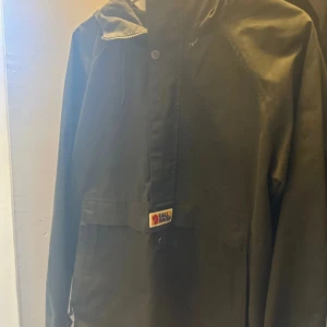 Anorak från Fjällräven M - Snygg svart vindjacka från Fjällräven i storlek M. Jackan har huva med snörning, klassisk logga på bröstet och svensk flagga på ärmen. Stängs med både dragkedja och knappar. Perfekt för dig som gillar stilren och funktionell design.