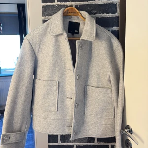 Ljusgrå overshirt från 157, storlek M - Stilren ljusgrå overshirt från 157 med klassisk krage, stora framfickor och knäppning framtill. Jackan har raka linjer och är tillverkad i ett mjukt material som ger en clean look. Perfekt för lager-på-lager och enkel att matcha.