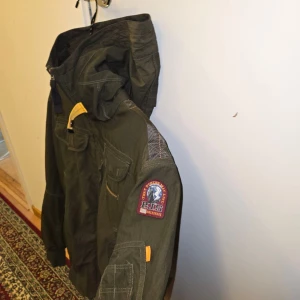 Grön vindjacka Parajumpers med huva - Snygg grön vindjacka från Parajumpers med flera fickor, huva och ikonisk gul detalj vid halsen. Jackan har patch på ärmen och dragkedja framtill. Perfekt för dig som gillar streetwear och funktionella plagg med coola detaljer.