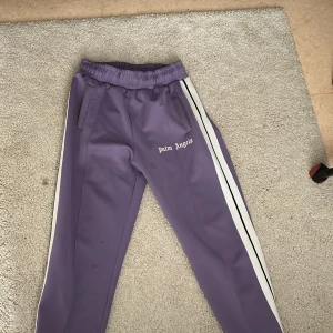 Lila Palm Angels trackpants - Snygga lila trackpants från Palm Angels med vita sidoränder och logga på benet. Byxorna har elastisk midja, raka ben och dragkedja vid benslut. Perfekta för en streetwear-look och tillverkade i glansig polyester.