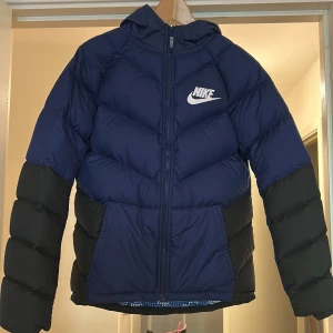 Nike blå och svart pufferjacka XL - Nike pufferjacka i blått och svart med huva och dragkedja. Jackan har ett stort Nike-tryck på bröstet, två fickor och coolt mönstrat innerfoder i blått och vitt. Perfekt för kalla dagar och riktigt snygg streetstil.