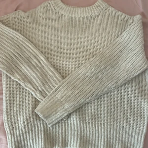 Beige stickad tröja från 157 - Mysig beige stickad tröja från 157 med grov ribbstickad struktur och rund hals. Tröjan har långärmade ärmar och en relaxed passform, perfekt för lager på lager under kyliga dagar. Enkel och stilren design som funkar till det mesta.