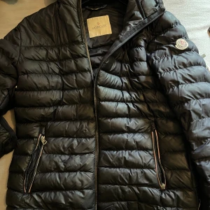 Svart Moncler pufferjacka med dragkedja - Snygg svart pufferjacka från Moncler med klassisk logga på ärmen. Jackan har horisontella sömmar, två fickor med dragkedja och detaljer i rött, vitt och blått vid fickorna. Perfekt för kyliga dagar och har en modern, sportig look.