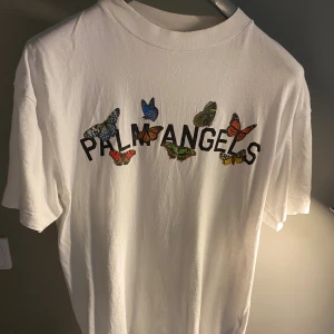 Palm Angels t-shirt - Säljer en vit t-shirt från Palm Angels med färgglada fjärilar och logotypen tryckt på bröstet. T-shirten har klassisk rund hals och korta ärmar. Perfekt för dig som gillar streetwear och vill sticka ut med snygga detaljer. Självklart äkta