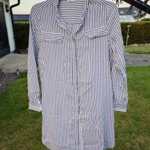 Blåvit randig lång skjorta från C - Snygg lång skjorta från C i storlek 36 med blå och vita vertikala ränder. Skjortan har knappar hela vägen, två bröstfickor med lock och klassisk krage. Långärmad och tillverkad i mjuk bomull, perfekt för en chill och stilren look.