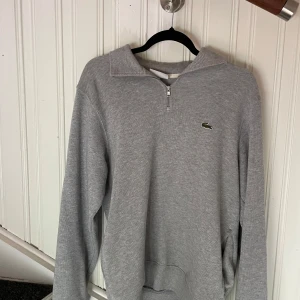 Grå half zip tröja från Lacoste - Grå långärmad tröja från Lacoste med klassisk krokodil-logga på bröstet. Tröjan har en snygg half zip-dragkedja vid halsen och är tillverkad i mjuk bomull. Perfekt för en chill och stilren look.