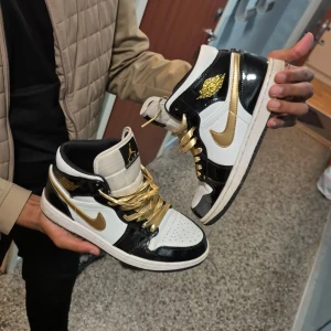 Nike Air Jordan 1 Gold/Black/White - Säljer ett par Nike Air Jordan 1 med svart, vit och guld färg. Skorna har glänsande svarta paneler, vita partier och metallicguld Swoosh samt logga. Guldiga skosnören och klassisk hög siluett. Perfekta för dig som vill sticka ut med en ikonisk sneaker.