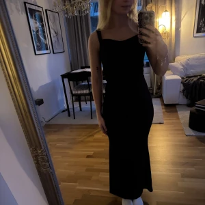 Svart långklänning med öppen rygg - Supersnygg svart långklänning med breda axelband och en riktigt snygg öppen rygg. Klänningen har en figurnära passform och stilren design som ger en elegant siluett.