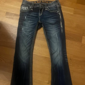 Rock Revival bootcut jeans med brodyr - Mörkblå low waisted Rock Revival bootcut jeans i storlek 27! Säljer dessa snygga jeans eftersom de tyvärr är lite för korta på mig. Nästan aldrig använda, köpte på vinted innan. Midjemått tvärs över: ca 36cm. Innerbenslängd: 72cm kontakta mig vid fler bilden eller frågor!! 💓