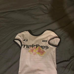 Vit Maldives t-shirt med blommor - Säljer en vit t-shirt från Maldives med svarta detaljer vid hals och ärmslut. Framsidan har tryck med text och två rosa-gula blommor. Kortärmad och med sportig vibe, perfekt för dig som gillar unik stil.