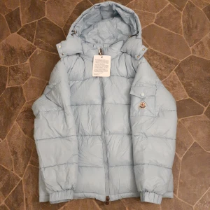Ljusblå Moncler dunjacka med huva - Snygg ljusblå dunjacka från Moncler med huva och klassisk logga på ärmen. Jackan har quiltad design, dragkedja framtill och en ficka med lock på ärmen. Perfekt för kalla dagar när du vill hålla stilen och samtidigt vara varm.