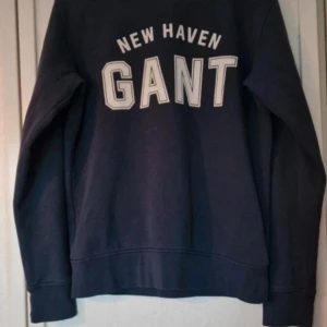 Mörkblå sweatshirt från Gant - Mörkblå sweatshirt från Gant med stor vit logga och texten 'New Haven Gant' på bröstet. Klassisk rund hals, långa ärmar och mjuk insida. 