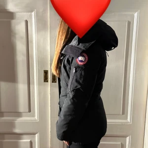 Svart dunjacka från Canada Goose 2XS - Säljer en svart dunjacka från Canada Goose i storlek 2XS. Jackan har huva, dragkedja och klassisk patch på ärmen. Perfekt för kalla dagar och har en snygg, figurnära passform. Fodrad och riktigt varm, med flera praktiska fickor och detaljer.