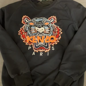 Svart Kenzo sweatshirt med tigerbrodyr - Svart sweatshirt från Kenzo med ikonisk tigerbrodyr i blått, orange och vitt framtill. Tröjan har rund hals, långa ärmar och ribbade muddar. Stort broderat Kenzo Paris-tryck i orange och vitt ger en riktigt fet streetkänsla.
