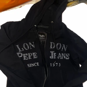 Svart hoodie från Pepe Jeans med strass - Xs/s den är figursydd så den sitter så fint på. Svart hoodie från Pepe Jeans med huva och dragkedja. Framsidan är dekorerad med texten 'London Pepe Jeans Since 1973' i glittrande strass. Tröjan har snörning i huvan och två fickor framtill. Perfekt för en cool och avslappnad stil.