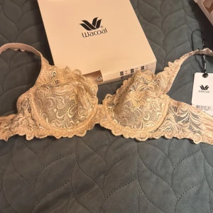 Beige bh från Wacoal - Supersnygg beige bh från Wacoal med blommigt spetsmönster och tunna axelband. Bh har vackra detaljer och transparent känsla, perfekt för att ge din outfit en extra touch. Justerbara axelband och hakknäppning bak för bästa passform.
